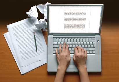 a pencil on a stack of written paper and lumps of crumpled paper, next to hands typing on a laptop computer with the screen showing a word processor program